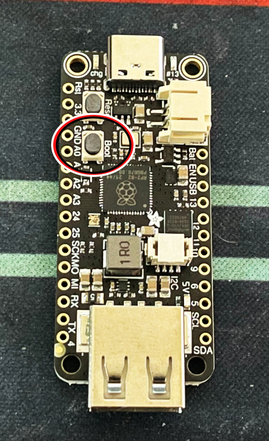 Una imagen de una Adafruit Feather RP2040. Un botón nombrado "Boot" esta circulado en rojo, blanco y negro.